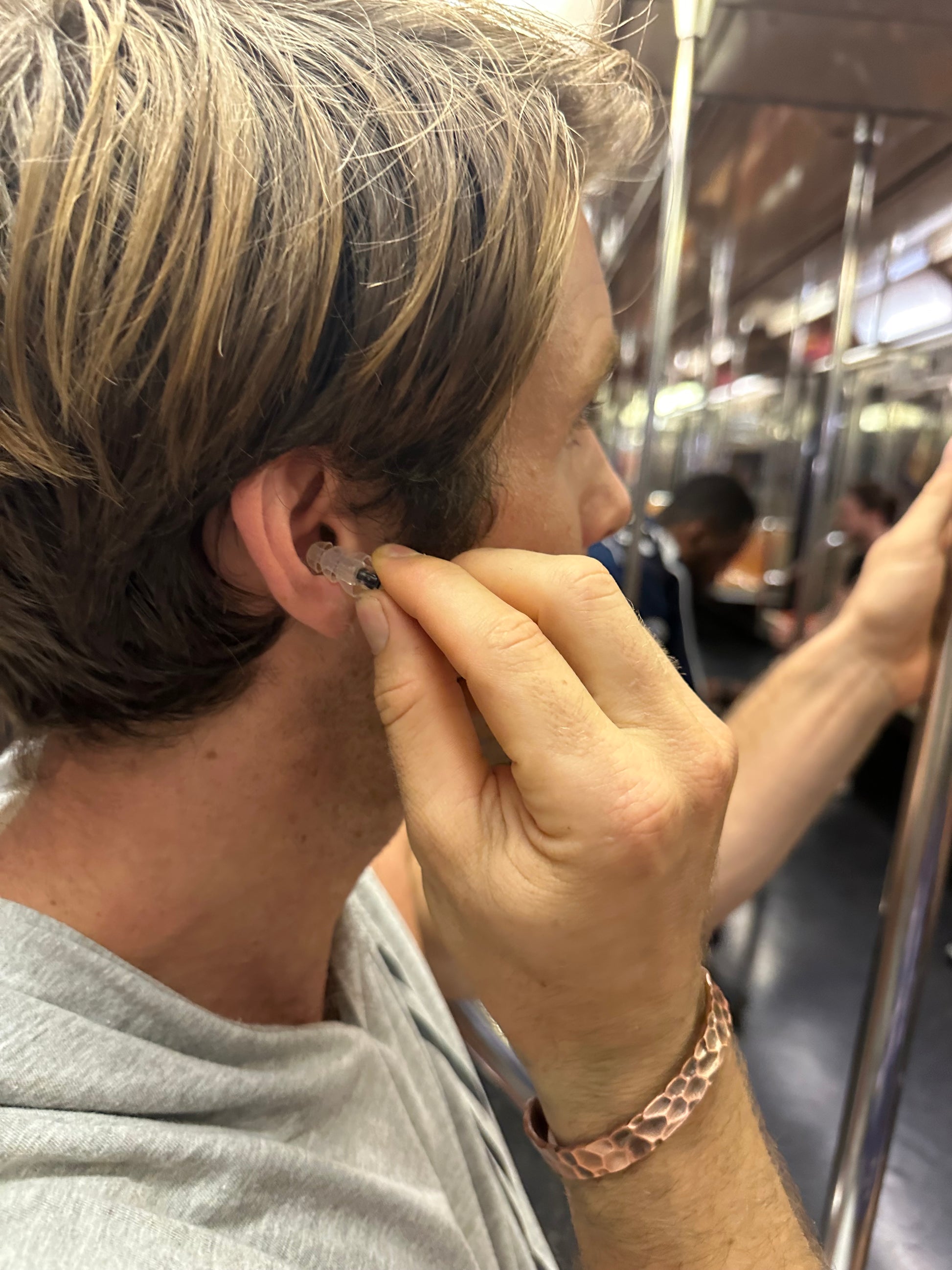 Ian Anderson inserting Orbis Earplugs into his ear while standing on a crowded subway, highlighting the earplugs' effectiveness in reducing noise in public transportation.